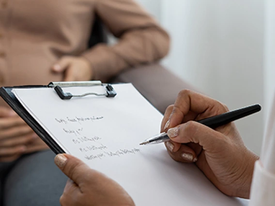 Doctor with clip board, taking notes on a pregnant patient who is seated in the backgroud.